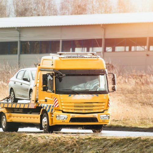 a roadside assistance service on a country road. A tow truck is carrying a car that has been involved in a traffic accident and has a broken engine